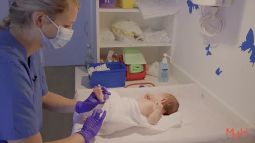 Frau mit blauem Kittel, Atemschutzmaske und Handschuhen wickelt einen Säugling.